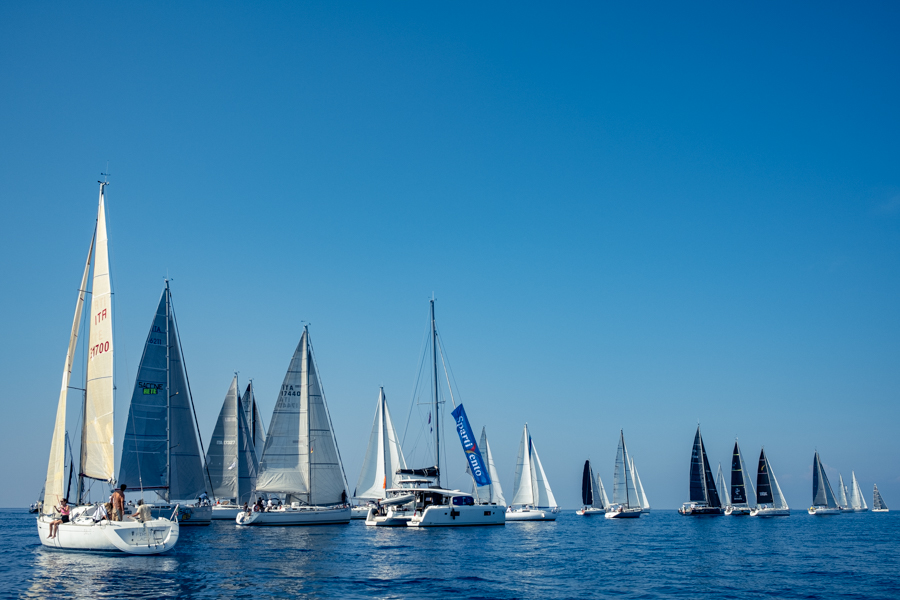 Barche a vela con vele aperte tagliano la linea di partenza durante la regata VELA Cup Sicilia 2023.